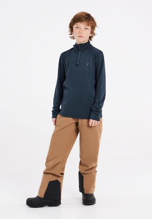 Dunkelblauer Pullover mit langen Ärmeln und halbem Reißverschlusskragen sowie Markenlogo; kombiniert mit beigen wasserdichten Hosen mit schwarzen Akzenten und robusten schwarzen Stiefeln.