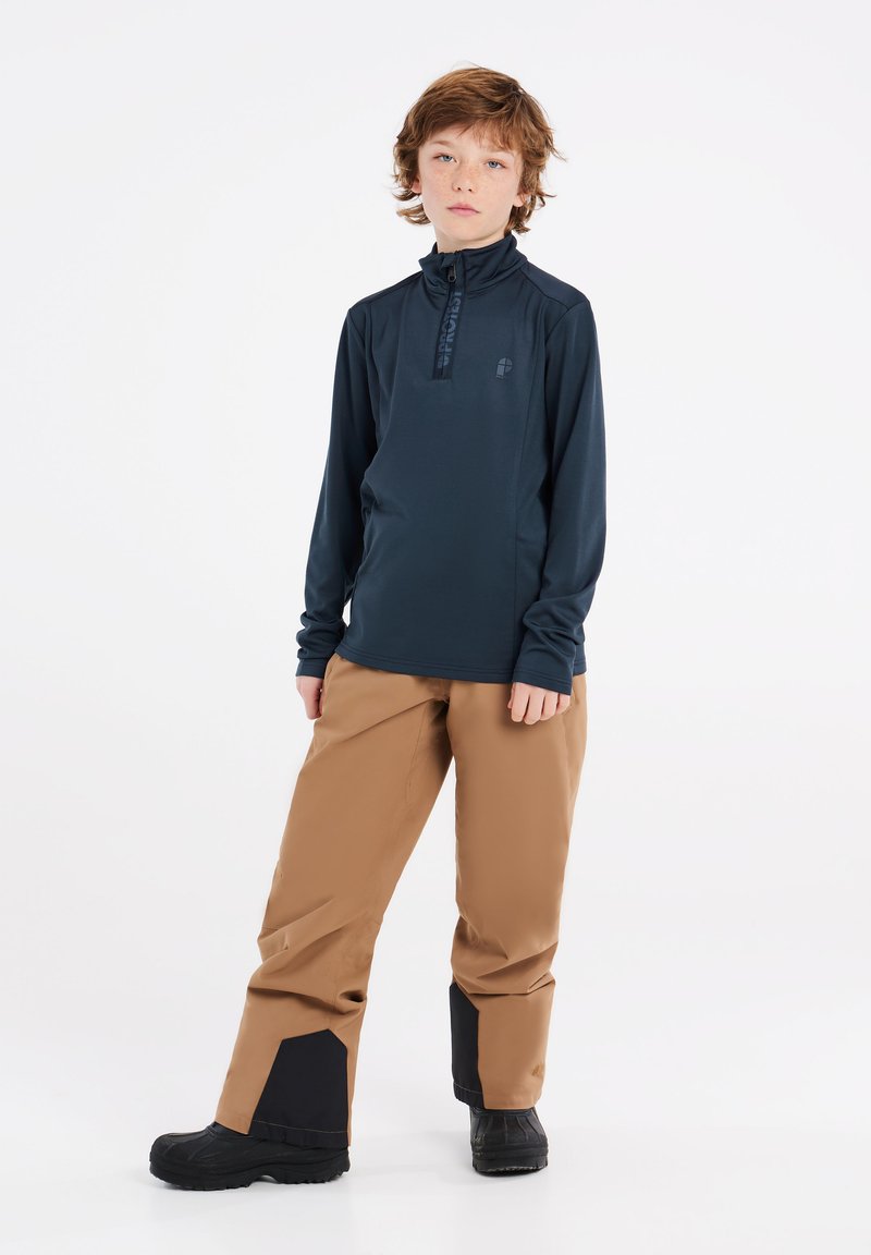 Langsleeve marineblauwe pullover met een halve rits en merkembleem; gecombineerd met tan waterdichte broeken met zwarte accenten en stevige zwarte laarzen.