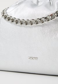 Sudrabaini ādas rokassoma ar savelkamu, aukliņu aizdari un sudraba ķēdes akcentu. Uz priekšpuses ir neliels DKNY logo.