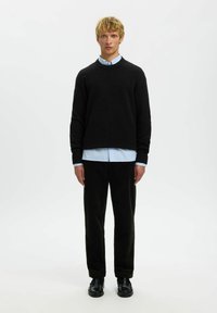 Noor blondide juustega noor inimene, kes kannab musta kampsunit helesinise särgi peal, musti pükse ja musti jalatseid, seismas puhta valge tausta ees.