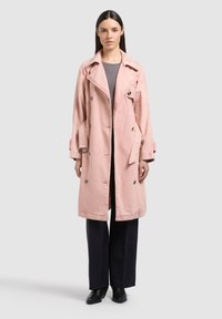 Donna in piedi che indossa un trench doppiopetto rosa chiaro, top grigio, pantaloni neri a gamba larga e stivaletti neri, su uno sfondo semplice.