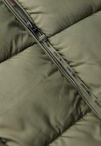 Olivgrön pufferjacka i tyg med dragkedjestängning. Syingarna skapar kviltade sektioner. Har en märkesdragkedjepull och texturerad foder.