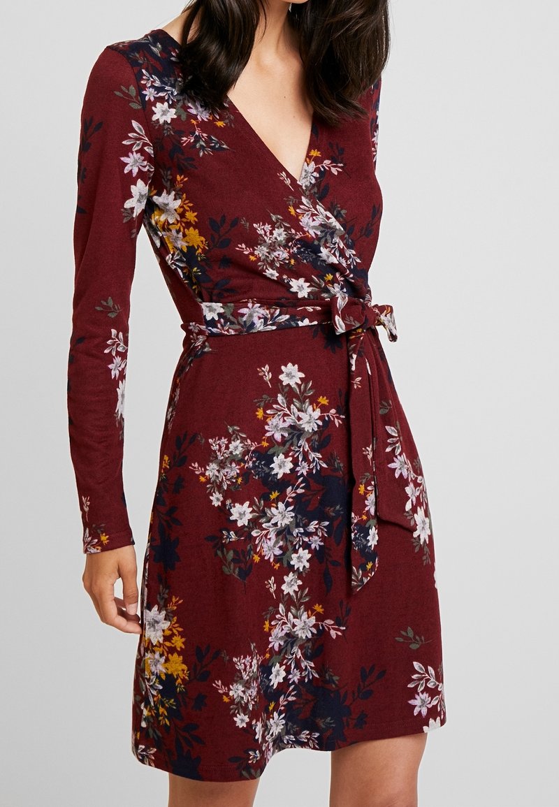 Vikklänning i bordeauxfärgad tyg med långa ärmar, prydd med ett blommönster i vitt, gult och mörkgrönt, med en knytning vid midjan.