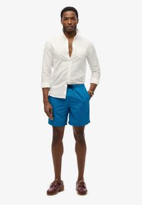 Man in een ongebotonneerd wit shirt, blauwe zwembroek met trekband, bruine leren boot schoenen en een horloge, staande tegen een witte achtergrond.