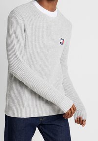 Hellgrauer gestrickter Pullover mit Rundhalsausschnitt, gerippten Bündchen und Saum, sowie einem kleinen Logopatch auf der Brust. Getragen über einem weißen Hemd.