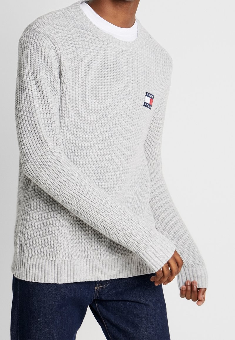 Hellgrauer gestrickter Pullover mit Rundhalsausschnitt, gerippten Bündchen und Saum, sowie einem kleinen Logopatch auf der Brust. Getragen über einem weißen Hemd.