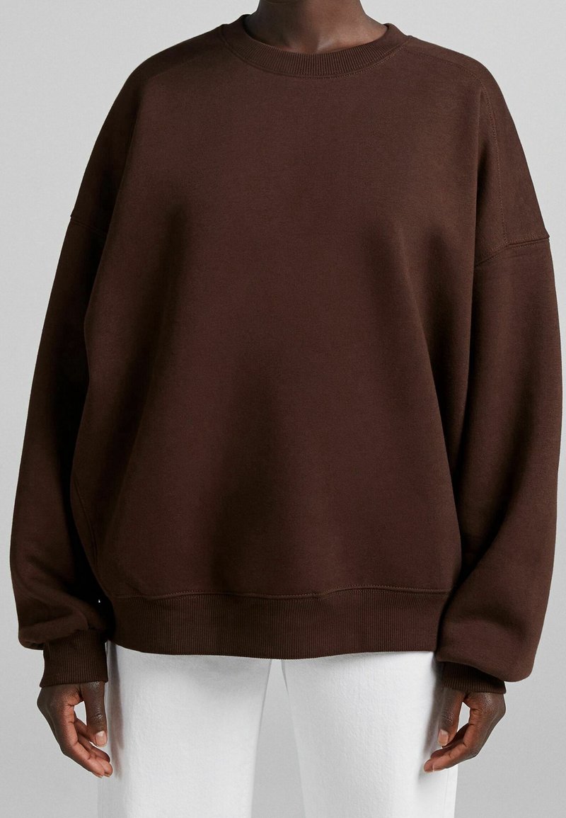 Personne portant un sweat-shirt marron foncé surdimensionné avec des poignets et un ourlet côtelés, associé à un pantalon blanc sur un fond uni.