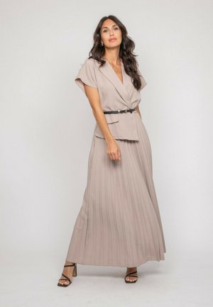 Femme portant un blazer beige à manches courtes ceinturé et une jupe maxi plissée assortie, debout devant un fond clair uni.