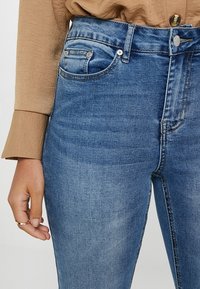 Jean de mezclilla azul de cintura alta con un acabado desgastado, que cuenta con cinco bolsillos, cierre de botón metálico y una silueta ajustada.