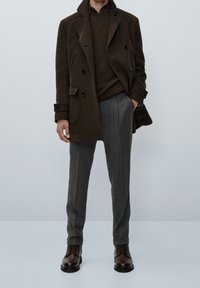 Cappotto di lana doppiopetto marrone sopra un maglione di maglia marrone scuro, abbinato a pantaloni grigio a fantasia e stivali di pelle marrone.