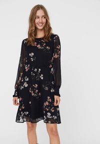 Robe noire à motifs floraux avec de longues manches transparentes, des poignets froncés et une taille ajustée. Elle présente un délicat motif floral en rose et gris.