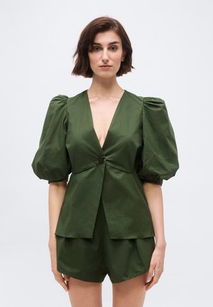 Kvinne med kort brunt hår iført en olivengrønn drakt med dyp V-hals, puffede korte ermer og matchende shorts.