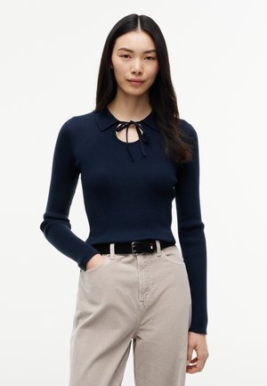 Kvinde med langt mørkt hår iført en marineblå ribstrikket sweater med krave og bindedetalje, beige bukser og sort bælte, står med den ene hånd i lommen.