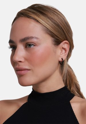 Top nero a coste con collo alto e design a spalla scoperta; la modella indossa un semplice orecchino ad anello nero, ha i capelli lisci e un'espressione neutra.
