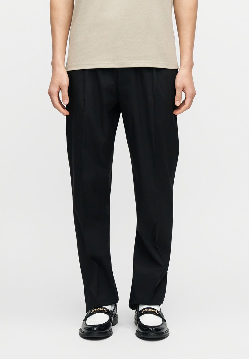 Μαύρο πλισέ παντελόνι με ευθεία γρ αμμή, ελαφρύ ύφασμα και διακριτική λάμψη, σε συνδυασμό με μαύρα και λευκά παπούτσια τύπου slip-on.