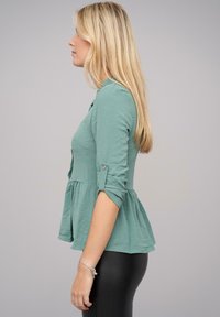 Top peplum verde teal in tessuto morbido e testurizzato. Presenta maniche arrotolate e un dettaglio con bottone. Abbinato a leggings neri.