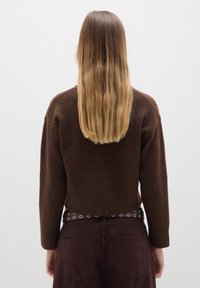 Suéter corto de lana marrón con dobladillo acanalado y mangas largas, combinado con pantalones marrones oscuros que cuentan con un cinturón con ojales de metal.