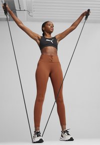 Sort sports-bh med hvidt logo, højtaljede brune leggings med snørredetalje, og sorte og hvide træningssko.