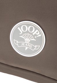 Hnedá textúrovaná taška z látky s okrúhlou striebornou logom s nápismi „JOOP!“ a „JEANS“, doplnená kvetinovým motifom a dekoračným rámom.