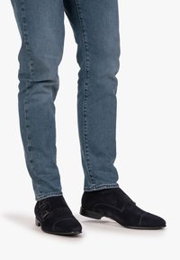 Suedeskor i marinblått med en slät yta, dubbel monk strap-spänne och rundad tådesign, tillsammans med åtsittande ljusblå jeans.