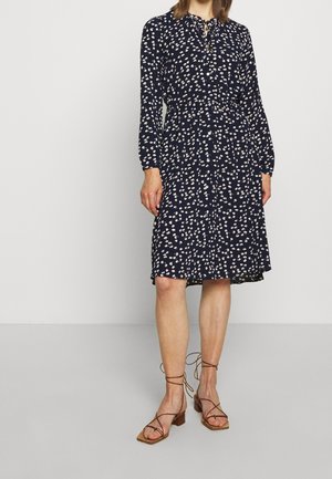 Femme portant une robe bleu marine mi-longue à petits motifs floraux blancs et des sandales à talons marron à lanières, debout devant un fond uni.