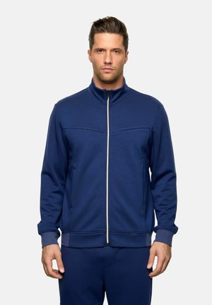 Hombre con cabello corto castaño, vistiendo una chaqueta azul marino con cremallera y pantalones a juego, de pie contra un fondo claro y liso.