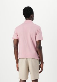 Ανοιχτό ροζ πόλο μπλούζα με κοντό γιακά, κοντά μανίκια και άνετη εφαρμογή, συνδυασμένη με μπεζ σορτσάκι. Λείος υφασμάτινος αφής, ελάχιστες λεπτομέρειες.