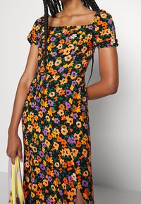 Abito floreale con scollatura quadrata, caratterizzato da uno sfondo nero, fiori arancioni, viola e gialli, e dettaglio con spacco laterale.