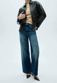 Schwarze lederne cropped Jacke mit braunen Nähten, beiger Pullover, weit geschnittene blaue Jeans und spitze schwarze Absätze. Clutch-Tasche wird vorne gehalten.