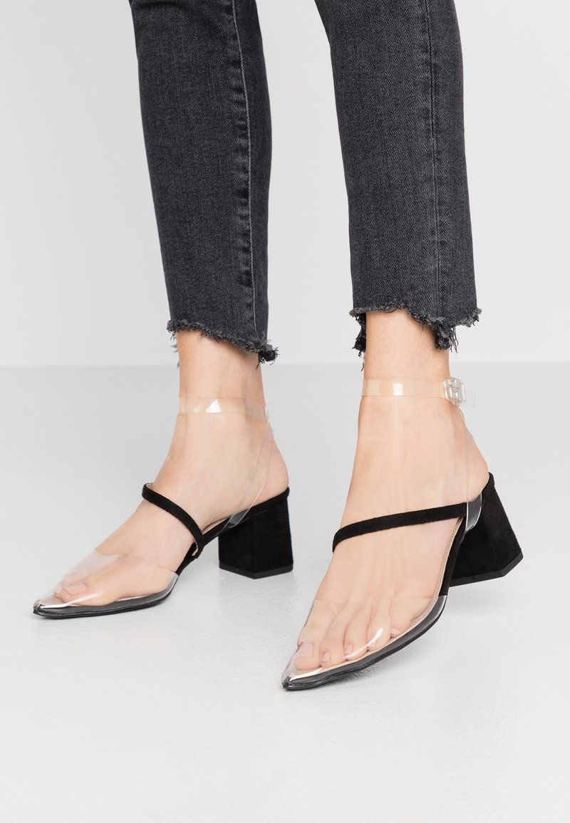 Pieds portant des sandales noires à talon bloc avec des brides transparentes et des bouts pointus, associées à un jean noir à ourlet effiloché.