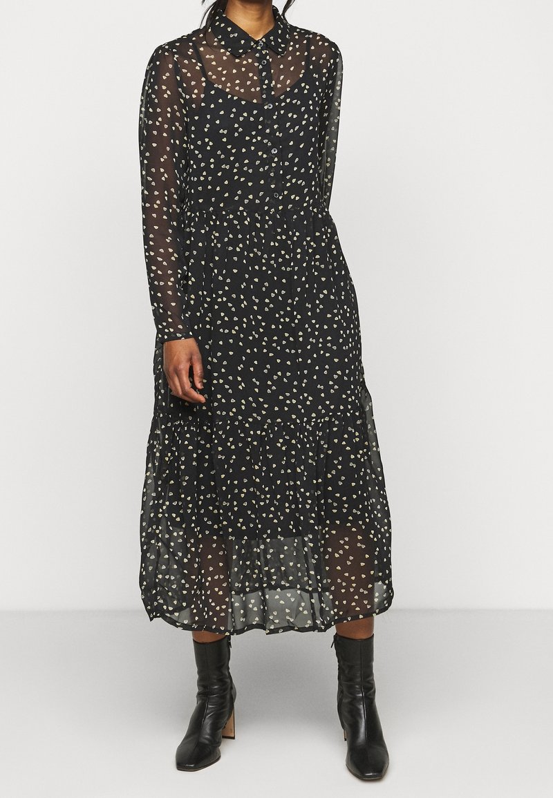 Femme portant une robe midi noire transparente à manches longues avec des petits motifs de cœurs blancs et des bottines noires.