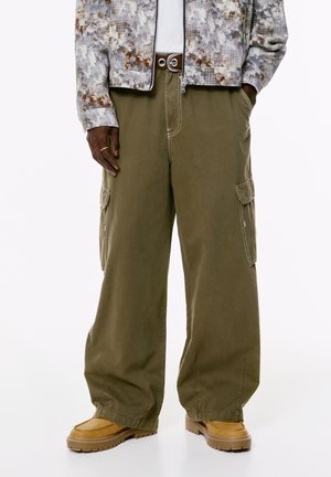 Persoană îmbrăcată în pantaloni cargo largi de culoare verde măsliniu, ghetuțe bej, un brâu negru, o bluză albă și o jachetă gri cu model, având mâinile în buzunare.