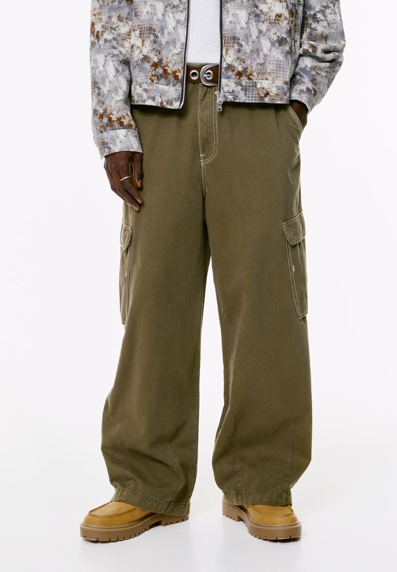 Persona che indossa pantaloni cargo larghi verde oliva, stivali beige, una cintura nera, una maglietta bianca e una giacca grigia con fantasia, con le mani in tasca.