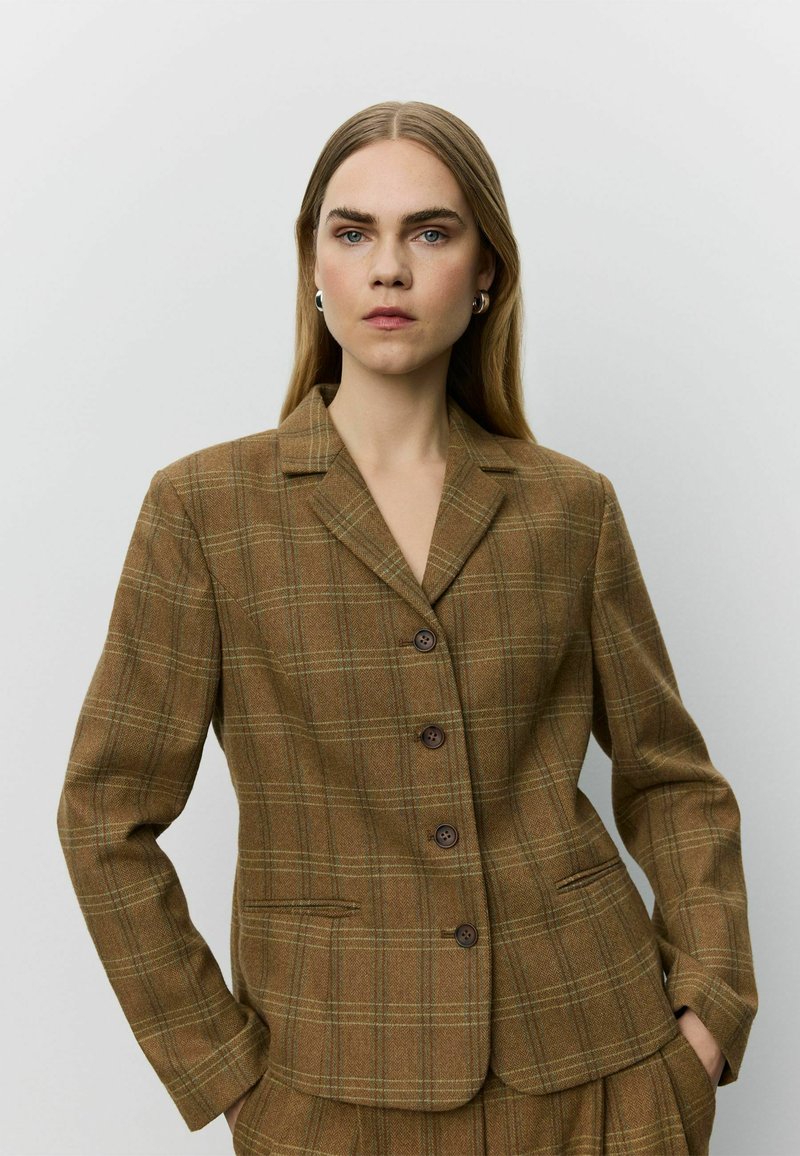 Femme aux longs cheveux blonds portant des boucles d'oreilles anneaux argentées et un blazer marron à carreaux boutonné, les mains dans les poches, sur un fond uni.