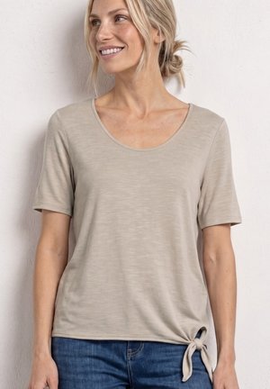 Kvinde med blondt hår i en lav knold iført en beige kortærmet top med en knude i siden og blå jeans, smilende og kigger mod højre.