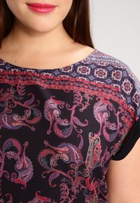 Sort mønstret top med et paisley-design i rødt, blåt og lilla. Inkluderer en rund halsudskæring og korte ærmer med en dekorativ kant.
