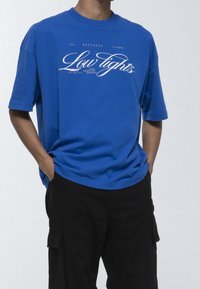 Modré bavlněné tričko s pohodlným střihem. Na přední straně se nachází bílý grafický potisk s nápisem "Low Lights", má krátké rukávy a kulatý výstřih.