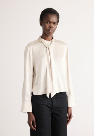 Beige Seidenbluse mit langen Ärmeln, einem gebundenen Kragen und ausgestellten Manschetten. Getragen mit schwarzen Hosen, die subtile glitzernde Akzente haben.
