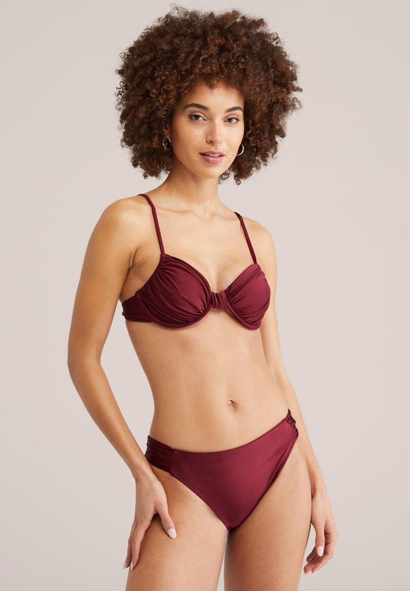 Femme aux cheveux bouclés portant un bikini bordeaux avec des détails froncés, se tenant contre un fond clair uni, posant avec une main sur la hanche.