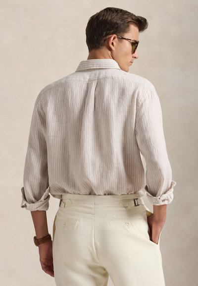 Ανοιχτόχρωμο ριγέ πουκάμισο με μακριά μανίκια και γιακά με κουμπιά, συνδυασμένο με μπεζ παντελόνια που διαθέτουν ρυθμιστικά πλαϊνά και πίσω τσέπες.