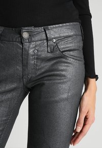 Un jean métallique noir présente une coupe slim, une texture brillante, un design à cinq poches et une fermeture à bouton à l'avant avec un accent métallique de marque.