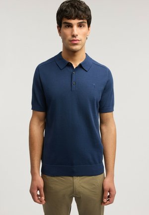 Mand med krøllet, mørkt hår står frontalt iført en mørkeblå kortærmet polo og beige bukser mod en ensfarvet baggrund.