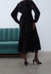 Czarna długa sukienka w kwiatowy wzór z dopasowaną talią i bufkami na rękawach, zestawiona z czarnymi sandałkami na paski, na tle zielononiebieskiej sofy.