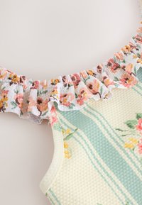 Vrh brez rokavov z cvetličnim vzorcem in naborki na izrezu, teksturirano tkanino v zelenih in rumenih črtah ter poudarki rožnate in oranžne barve.