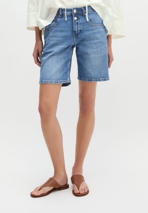 Persoon draagt mid-lange blauwe denimshorts, een wit los topje en bruine open-toe platte sandalen, staand tegen een effen achtergrond.
