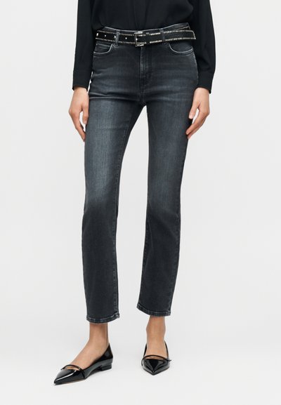 Dunkelgraue Jeans mit schmaler Passform, vorderen Taschen, einem schwarzen, mit Nieten besetzten Gürtel und einer verkürzten Länge, kombiniert mit spitzen schwarzen Ballerinas.