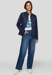 Frau in einer dunkelblauen Steppjacke über einem weißen T-Shirt mit Grafik, weitem blauen Jeans und weißen Sneakern, steht mit einer Hand in der Tasche.