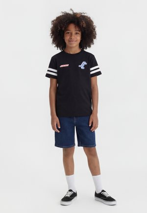 Παιδί με κατσαρά μαλλιά που φοράει μαύρο μπλουζάκι με σχέδιο, τζιν σορτσάκι, άσπρες κάλτσες και μαύρα αθλητικά παπούτσια, στέκεται μπροστά σε απλό φόντο.