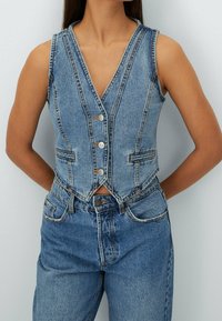 Ärmlös blå denimväst med framknappar, bärs över blå denimjeans, personens armar bakom ryggen, mot en ljus enfärgad bakgrund.