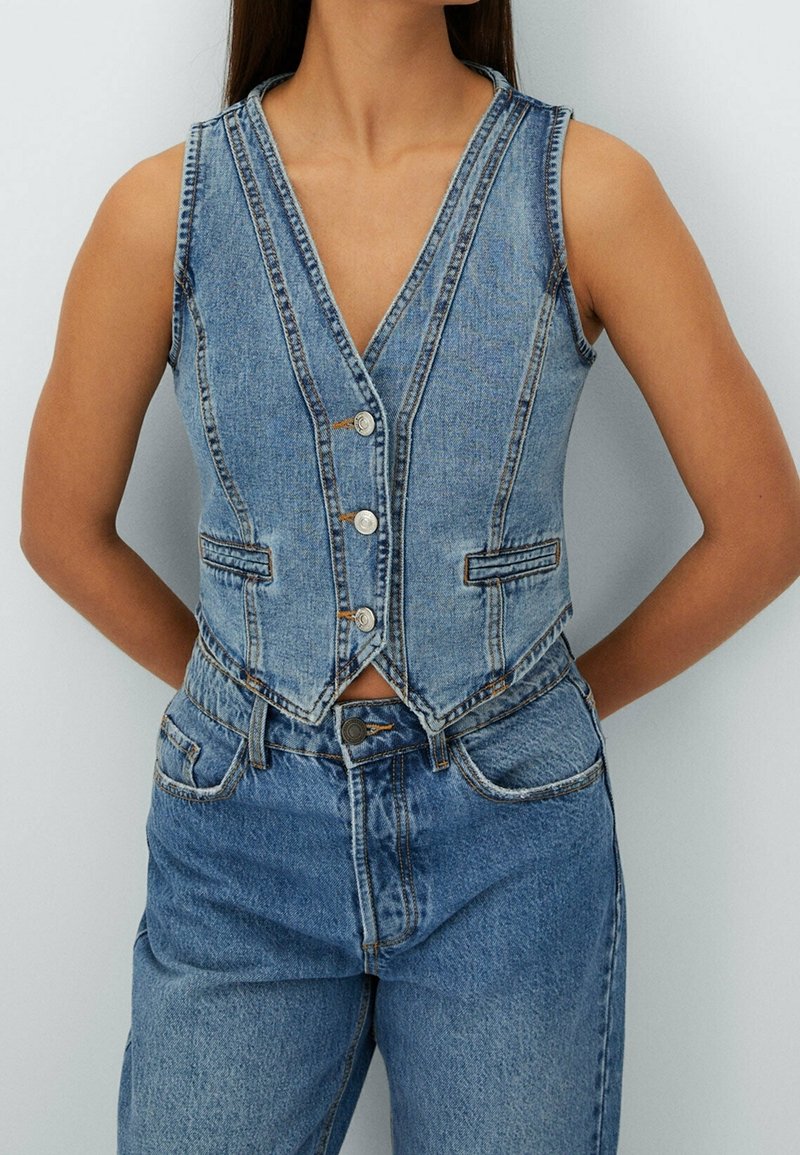 Ärmlös blå denimväst med framknappar, bärs över blå denimjeans, personens armar bakom ryggen, mot en ljus enfärgad bakgrund.